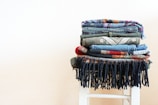 A neatly folded stack of various colored and patterned textiles is placed on a white stool. The textures range from denim to knits, featuring an assortment of earthy and vibrant colors. The bottom textile has black fringes that hang down over the stool.