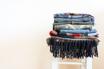 A neatly folded stack of various colored and patterned textiles is placed on a white stool. The textures range from denim to knits, featuring an assortment of earthy and vibrant colors. The bottom textile has black fringes that hang down over the stool.