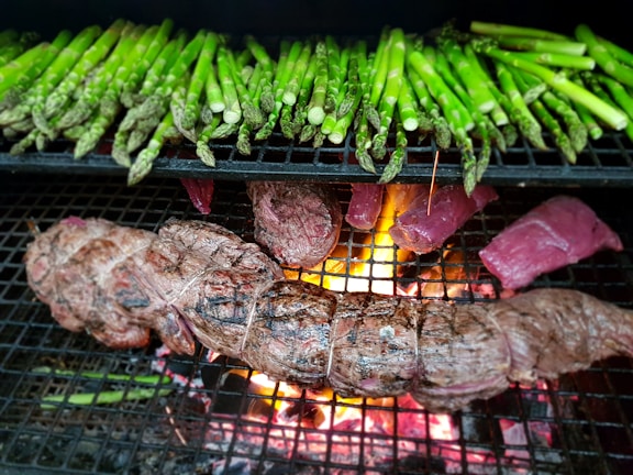 A grill loaded with green asparagus spears and various cuts of meat, including a tied roast and steaks. The flames and glowing embers from the fire beneath the grill are visible, adding heat and a vibrant orange glow to the scene.
