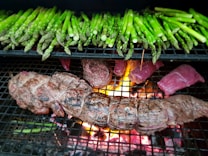 A grill loaded with green asparagus spears and various cuts of meat, including a tied roast and steaks. The flames and glowing embers from the fire beneath the grill are visible, adding heat and a vibrant orange glow to the scene.