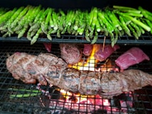 A grill loaded with green asparagus spears and various cuts of meat, including a tied roast and steaks. The flames and glowing embers from the fire beneath the grill are visible, adding heat and a vibrant orange glow to the scene.