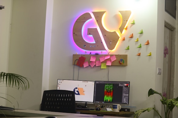 An office workspace with a modern decor theme. The wall displays a large wooden emblem with illuminated neon lights in purple and yellow. Below this emblem is a small shelf adorned with vibrant origami butterflies in orange, yellow, and green. Several colorful sticky notes are attached to the wooden board. A desk has two computer monitors displaying digital content, with a black mesh chair in front. There are indoor plants in the background, adding a touch of greenery.
