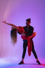 A man and a woman are captured in a dynamic dance pose, illuminated by vibrant purples and blues. The woman is wearing a red dress, gracefully arched backward with long, flowing hair cascading down.