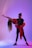 A man and a woman are captured in a dynamic dance pose, illuminated by vibrant purples and blues. The woman is wearing a red dress, gracefully arched backward with long, flowing hair cascading down.