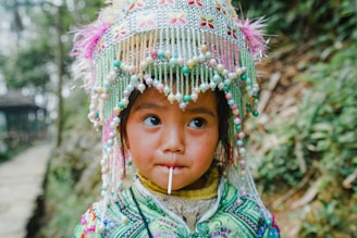 A young child wearing an ornate headdress decorated with colorful beads and embroidery is holding a lollipop. The background shows a blurred greenery and pathway, hinting at a natural outdoor setting.