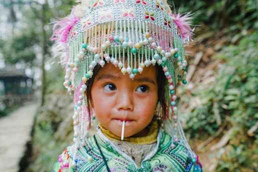 A young child wearing an ornate headdress decorated with colorful beads and embroidery is holding a lollipop. The background shows a blurred greenery and pathway, hinting at a natural outdoor setting.