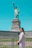 A smiling young woman standing proudly in front of the Statue of Liberty on a sunny day.