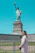 A smiling young woman standing proudly in front of the Statue of Liberty on a sunny day.