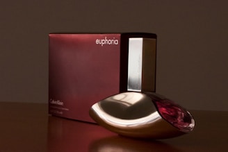 A bottle of Calvin Klein's Euphoria perfume is placed in front of its packaging. The bottle is sleek with a unique design and features metallic and transparent accents. The box is a deep burgundy color with minimalistic white text.