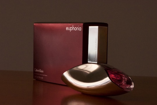 A bottle of Calvin Klein's Euphoria perfume is placed in front of its packaging. The bottle is sleek with a unique design and features metallic and transparent accents. The box is a deep burgundy color with minimalistic white text.