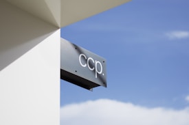 A metallic sign with the letters 'ccp.' is attached to a white wall. The sign is sleek and minimalistic, and the background consists of a clear blue sky with a few white clouds.