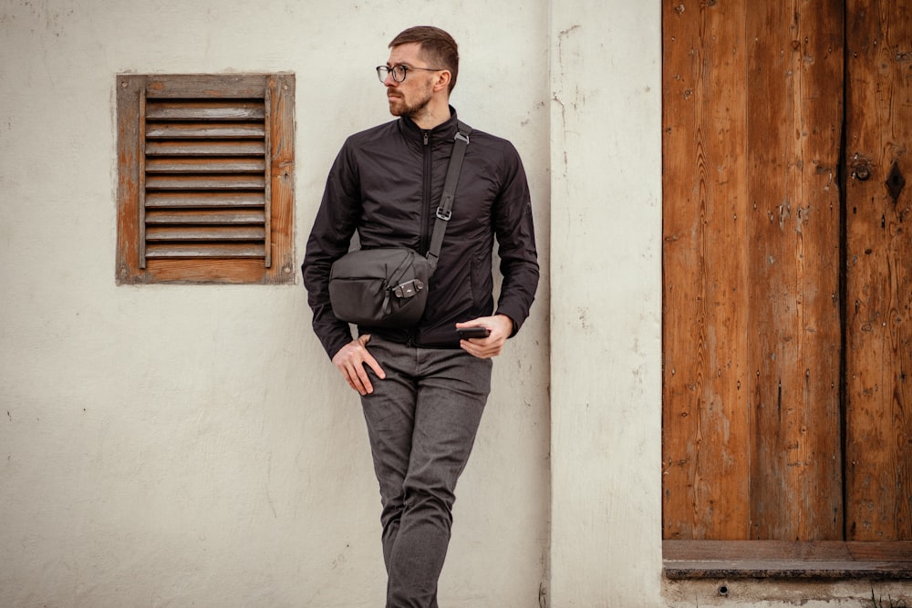 man wearing black zip-up jacket and black pants leaning on white painted wall