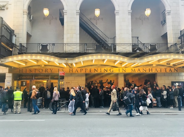 A crowded sidewalk in front of a theater with an illuminated marquee that reads 'HISTORY IS HAPPENING IN MANHATTAN'. The theater is adorned with large arched windows and intricately designed railings. Numerous people stand in line or walk past, some engaging in conversation.