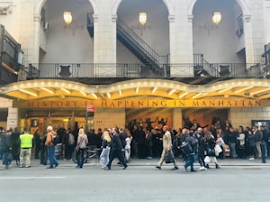 A crowded sidewalk in front of a theater with an illuminated marquee that reads 'HISTORY IS HAPPENING IN MANHATTAN'. The theater is adorned with large arched windows and intricately designed railings. Numerous people stand in line or walk past, some engaging in conversation.