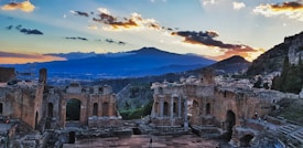 Ancient ruins with arches and columns are set against a backdrop of a mountainous landscape. The skyline features a dramatic sunset with vibrant clouds, casting warm light on the scene. The view offers a sense of historical grandeur and natural beauty, with distant buildings nestled on hillsides.
