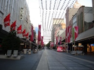 A city street scene with tram tracks running through the center. Buildings line both sides, adorned with red sale banners. A series of decorative elements hang across the street. The area is devoid of people, giving a calm feel. Retail store signs like MYER and Sussan are visible, suggesting a commercial district.
