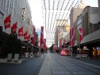 A city street scene with tram tracks running through the center. Buildings line both sides, adorned with red sale banners. A series of decorative elements hang across the street. The area is devoid of people, giving a calm feel. Retail store signs like MYER and Sussan are visible, suggesting a commercial district.