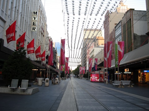 A city street scene with tram tracks running through the center. Buildings line both sides, adorned with red sale banners. A series of decorative elements hang across the street. The area is devoid of people, giving a calm feel. Retail store signs like MYER and Sussan are visible, suggesting a commercial district.