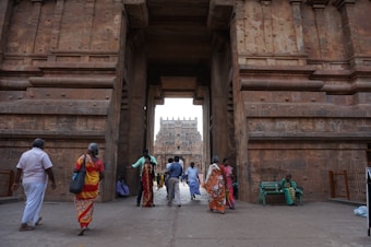 A group of people is walking through an ancient stone gateway, adorned with intricate carvings. The massive pillars frame the entrance to a temple complex visible in the distance. Some people are dressed in traditional attire, indicating a culturally significant location.