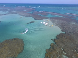 Aerial view of a coastal area with crystal-clear turquoise water and visible coral formations. Several boats and people are gathered on a sandy patch amidst the water, suggesting a recreational or tourist activity. A jet ski is creating a wake in the water nearby.