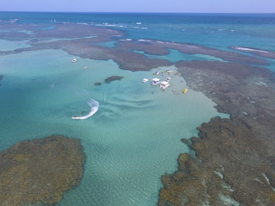 Aerial view of a coastal area with crystal-clear turquoise water and visible coral formations. Several boats and people are gathered on a sandy patch amidst the water, suggesting a recreational or tourist activity. A jet ski is creating a wake in the water nearby.