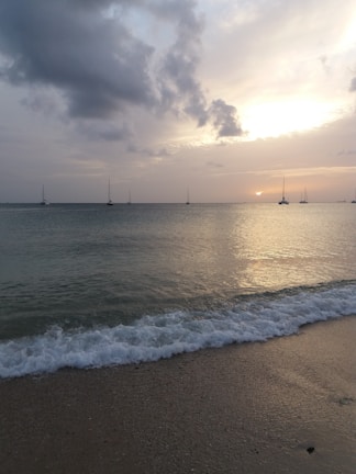 Golden hour light bathing a serene beach with gentle waves and distant sailboats.