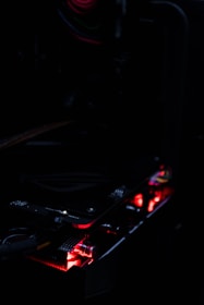A dark image focusing on a computer component, possibly a graphics card, with glowing red LED lights. The environment is dimly-lit, accentuating the red and subtle highlights from the hardware.