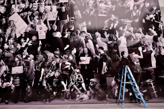 A monochrome collage of a mass gathering, likely a protest or rally, featuring diverse individuals holding various signs with messages. Prominent signs include feminist symbols and political statements. There is a mix of expressions, with most people appearing engaged or passionate. A blue ladder stands out in the foreground, contrasting against the black-and-white composition.
