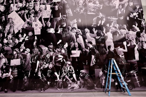 A monochrome collage of a mass gathering, likely a protest or rally, featuring diverse individuals holding various signs with messages. Prominent signs include feminist symbols and political statements. There is a mix of expressions, with most people appearing engaged or passionate. A blue ladder stands out in the foreground, contrasting against the black-and-white composition.
