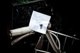 A handwritten note is attached to the handlebars of a bicycle. The note is secured with a small pin and contains some cursive writing. The bicycle handle is white with a vintage appearance, and there is a wire basket partially visible in the foreground. The background is dark, creating a contrast that highlights the note and the bicycle details.