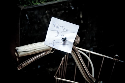 A handwritten note is attached to the handlebars of a bicycle. The note is secured with a small pin and contains some cursive writing. The bicycle handle is white with a vintage appearance, and there is a wire basket partially visible in the foreground. The background is dark, creating a contrast that highlights the note and the bicycle details.