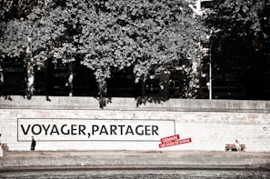 A riverside walkway features a large mural on a wall with the words 'VOYAGER, PARTAGER' and a smaller red sign beneath it. A person dressed in dark clothing is seen walking alone in front of the mural. Nearby, another person sits on a bench, enjoying the surroundings. Lush trees provide a canopy above the walkway.
