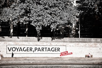 A riverside walkway features a large mural on a wall with the words 'VOYAGER, PARTAGER' and a smaller red sign beneath it. A person dressed in dark clothing is seen walking alone in front of the mural. Nearby, another person sits on a bench, enjoying the surroundings. Lush trees provide a canopy above the walkway.