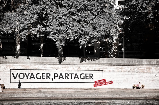 A riverside walkway features a large mural on a wall with the words 'VOYAGER, PARTAGER' and a smaller red sign beneath it. A person dressed in dark clothing is seen walking alone in front of the mural. Nearby, another person sits on a bench, enjoying the surroundings. Lush trees provide a canopy above the walkway.
