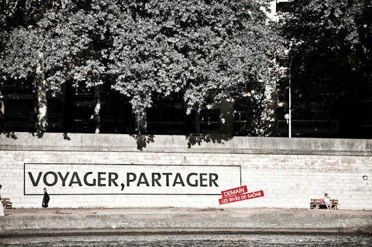 A riverside walkway features a large mural on a wall with the words 'VOYAGER, PARTAGER' and a smaller red sign beneath it. A person dressed in dark clothing is seen walking alone in front of the mural. Nearby, another person sits on a bench, enjoying the surroundings. Lush trees provide a canopy above the walkway.