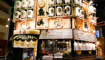 A brightly lit storefront with a traditional Japanese aesthetic featuring numerous white paper lanterns decorated with floral designs. The building has prominent wooden accents and Japanese characters displayed prominently on the facade. Spotlights are strategically placed, enhancing the overall warm and inviting atmosphere of a Japanese restaurant.
