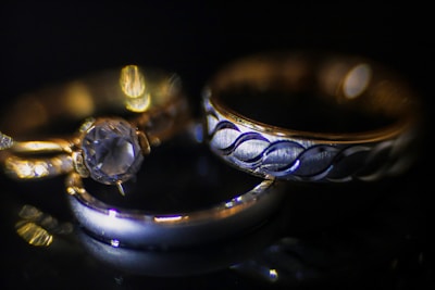 A close-up view of three rings placed on a dark surface. One of the rings features a large, sparkling gemstone, while the others display intricate metal patterns. The lighting creates a dramatic and elegant effect, highlighting the metallic shine and gemstone clarity.