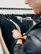 A person is browsing through a clothing rack filled with black and white garments. The focus is on the person's hands, one of which has a watch on the wrist. The garments are neatly hung on black hangers.