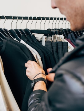 A person is browsing through a clothing rack filled with black and white garments. The focus is on the person's hands, one of which has a watch on the wrist. The garments are neatly hung on black hangers.