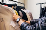 A person browses through a rack of clothing in a store, holding up a white T-shirt with text that reads 'NO FUN.' The clothes are hung neatly on black hangers and include a variety of colors from white to darker shades. The person is wearing a black watch and a dark, padded jacket.