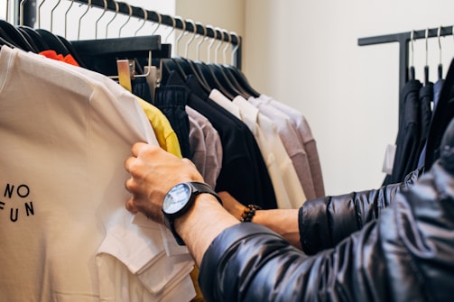 A person browses through a rack of clothing in a store, holding up a white T-shirt with text that reads 'NO FUN.' The clothes are hung neatly on black hangers and include a variety of colors from white to darker shades. The person is wearing a black watch and a dark, padded jacket.