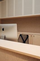 A modern office setup features a sleek computer sitting atop a wooden desk. The background displays perforated white panels and light-colored cabinets. A black mesh office chair is partially visible, contributing to a minimalistic and organized feel.