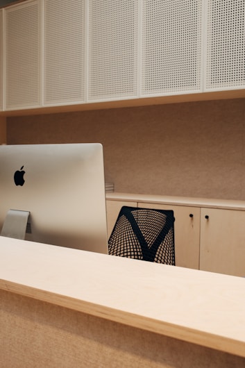 A modern office setup features a sleek computer sitting atop a wooden desk. The background displays perforated white panels and light-colored cabinets. A black mesh office chair is partially visible, contributing to a minimalistic and organized feel.