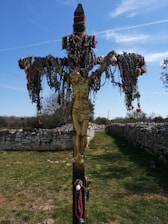 A crucifix adorned with numerous colorful rosaries and beads is set in an outdoor stone pathway. The sunny sky and sparse trees in the background add to the serene atmosphere.