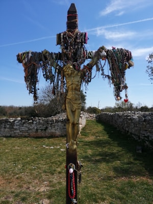 A crucifix adorned with numerous colorful rosaries and beads is set in an outdoor stone pathway. The sunny sky and sparse trees in the background add to the serene atmosphere.