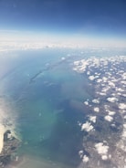 An aerial view captures a vast expanse of ocean, transitioning into land. The image features scattered white clouds floating over the blue-green waters of the sea, with visible patterns, possibly indicating underwater terrain or currents. The coastline shows areas of urban development and natural landscapes merging smoothly into the sea.