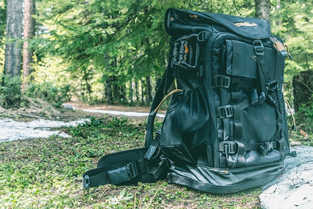 A large black backpack sits on a forest floor covered with green foliage. The backpack has multiple straps and compartments, suggesting it's designed for outdoor or hiking activities. Surrounding the backpack, lush green trees and dappled sunlight create a natural and serene setting.