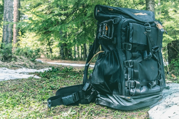 A large black backpack sits on a forest floor covered with green foliage. The backpack has multiple straps and compartments, suggesting it's designed for outdoor or hiking activities. Surrounding the backpack, lush green trees and dappled sunlight create a natural and serene setting.