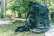 A large black backpack sits on a forest floor covered with green foliage. The backpack has multiple straps and compartments, suggesting it's designed for outdoor or hiking activities. Surrounding the backpack, lush green trees and dappled sunlight create a natural and serene setting.