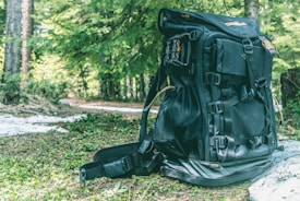 A large black backpack sits on a forest floor covered with green foliage. The backpack has multiple straps and compartments, suggesting it's designed for outdoor or hiking activities. Surrounding the backpack, lush green trees and dappled sunlight create a natural and serene setting.